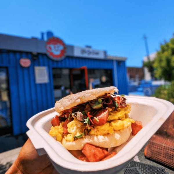 A breakfast sandwich with scrambled eggs and bacon in a takeout container, with Cafe Da Fonk in the background.