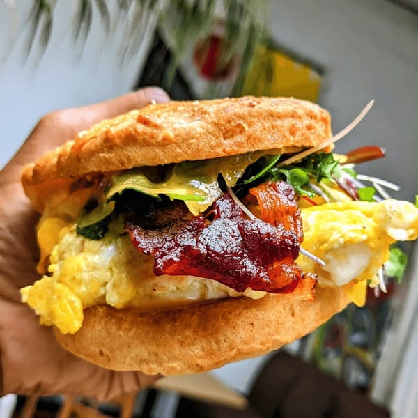 A hand holds a Cafe Da Fonk breakfast sandwich with bacon, scrambled eggs, and greens.