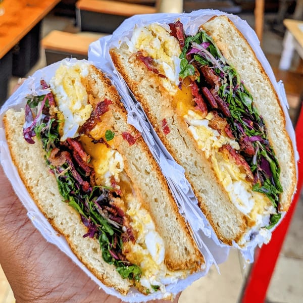 A hand holds a Cafe Da Fonk breakfast sandwich, cut in half, revealing egg, bacon, and greens.